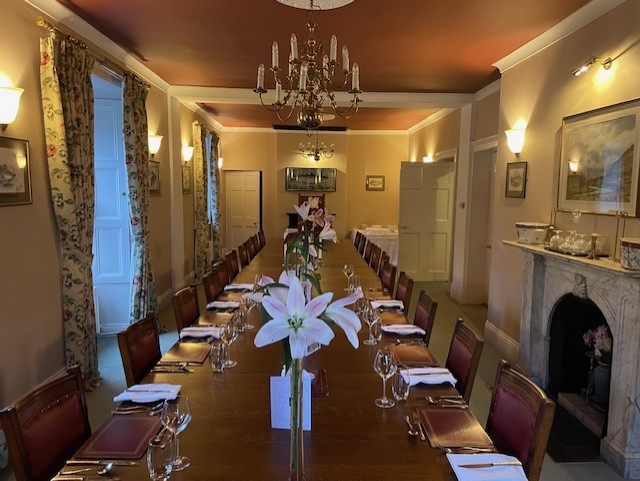 Een lange eettafel gedekt voor een formele maaltijd loopt door het midden van een elegante kamer met kroonluchters, stoelen met rode kussens, witte servetten, glaswerk en een marmeren open haard. Grote witte lelies in vazen staan op tafel; bloemige gordijnen omlijsten hoge ramen. | Echt Ierland Vakanties