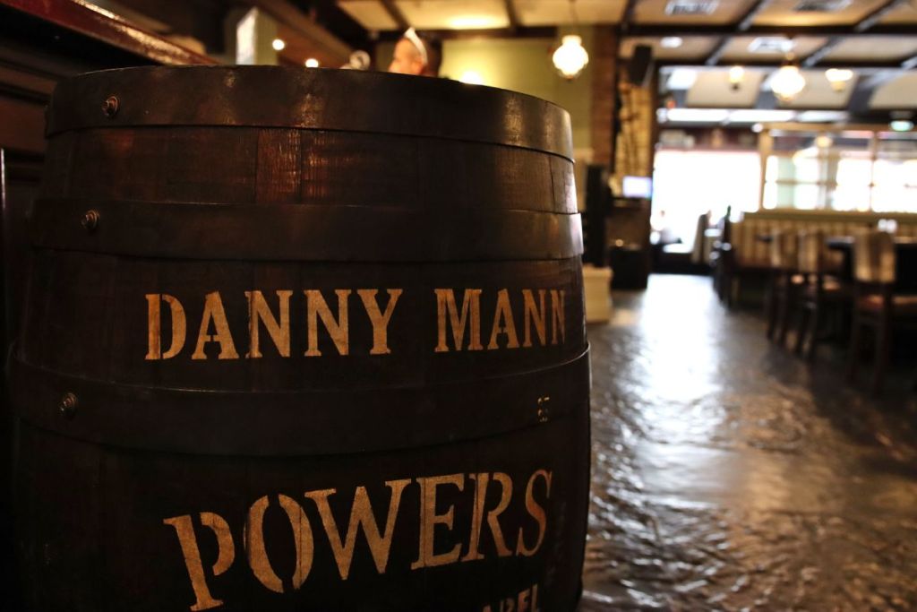 Een groot houten vat met de woorden DANNY MANN POWERS in goud geschilderd staat op de voorgrond van Brook Lodge Hotel, Killarney-een warm verlichte, ruime pub of restaurant met houten meubilair en een vloer met stenen textuur. Lege tafels vullen de achtergrond. | Echt Ierland Vakanties