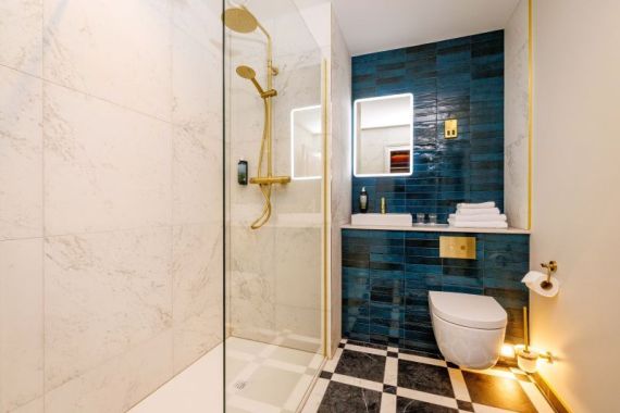Moderne badkamer met een glazen douche met gouden armaturen, marmeren tegelwanden en een donkerblauwe betegelde accentmuur achter een zwevende wastafel en een wandtoilet. Handdoeken en toiletartikelen zijn netjes gerangschikt; op de vloer liggen zwart-wit geblokte tegels. | Echt Ierland Vakanties