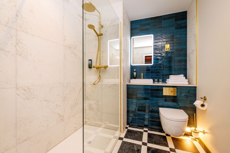 Moderne badkamer met een glazen douche met gouden armaturen, marmeren tegelwanden en een donkerblauwe betegelde accentmuur achter een zwevende wastafel en een wandtoilet. Handdoeken en toiletartikelen zijn netjes gerangschikt; op de vloer liggen zwart-wit geblokte tegels. | Echt Ierland Vakanties