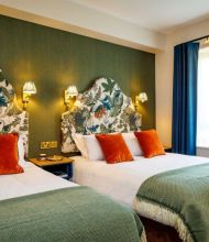 Een gezellige kamer in dit charmante Boutique-Hotel im Herzen von Killarney heeft twee tweepersoonsbedden met wit linnengoed, groene spreien en oranje kussens. Bloemige hoofdeinden, warme kandelaars, een blauw raam met gordijnen en een nachtkastje dragen bij aan de uitnodigende sfeer. | Echt Ierland Vakanties