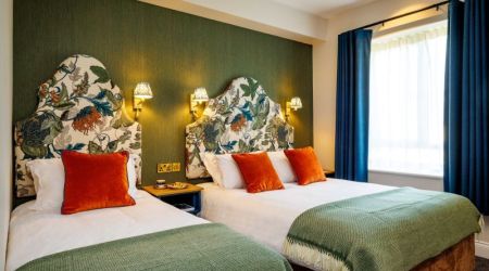 Een gezellige kamer in dit charmante Boutique-Hotel im Herzen von Killarney heeft twee tweepersoonsbedden met wit linnengoed, groene spreien en oranje kussens. Bloemige hoofdeinden, warme kandelaars, een blauw raam met gordijnen en een nachtkastje dragen bij aan de uitnodigende sfeer. | Echt Ierland Vakanties