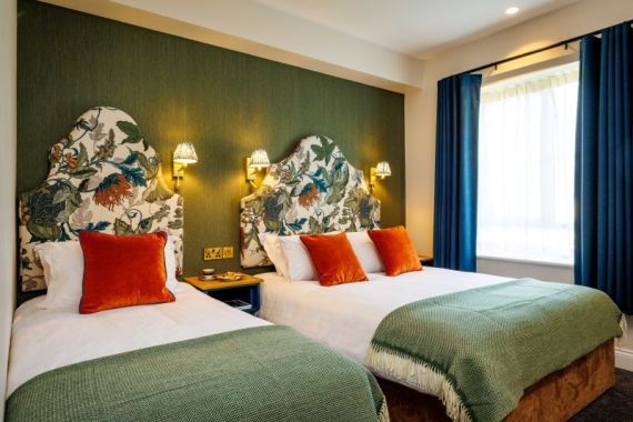 Een gezellige kamer in dit charmante Boutique-Hotel im Herzen von Killarney heeft twee tweepersoonsbedden met wit linnengoed, groene spreien en oranje kussens. Bloemige hoofdeinden, warme kandelaars, een blauw raam met gordijnen en een nachtkastje dragen bij aan de uitnodigende sfeer. | Echt Ierland Vakanties