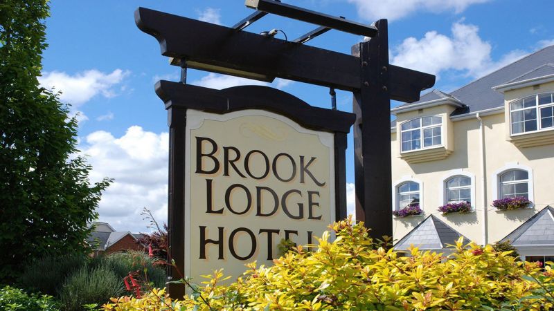 Een groot bord met de tekst Brook Lodge Hotel staat tussen geelgroene struiken. Op de achtergrond, bij Castlewellan, is een crèmekleurig gebouw met witte bies, grijs dak en bloembakken te zien onder een helderblauwe lucht met verspreide wolken. | Echt Ierland Vakanties