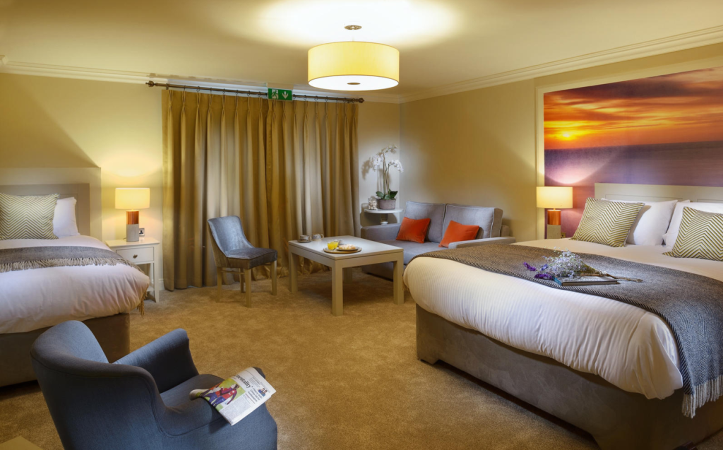 Een ruime hotelkamer met twee tweepersoonsbedden, beige muren, warme verlichting, een vloerbedekking, grijze leunstoelen, een tafel met versnaperingen, grote bruine gordijnen en een foto van een zonsondergang aan de muur. Accentkussens met oranje en groene patronen voegen kleur toe. | Echt Ierland Vakanties