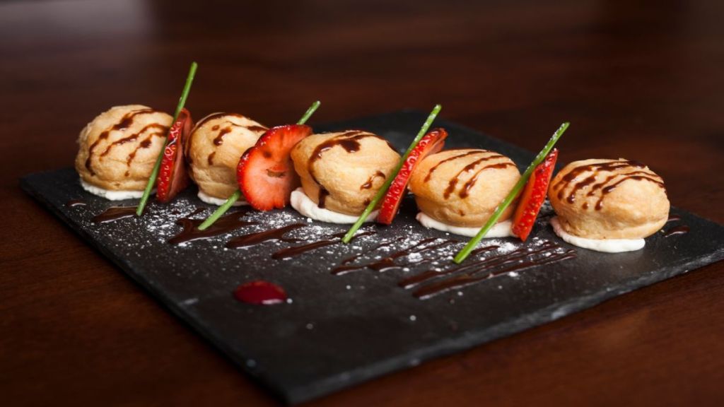 Op een bord van leisteen liggen vijf soesjes met chocolade eroverheen, elk op een toefje room. Plakjes aardbei en groene garnering liggen tussen de gebakjes, met poedersuiker en sausaccenten op het bord. | Echt Ierland Vakanties