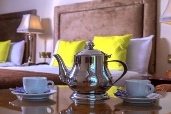 Een zilveren theepot en twee witte theekopjes met schotels staan op een glazen tafel in Hillyard House Hotel. Daarachter creëren twee knusse bedden met bruine hoofdeinden, gele kussens en warme lampen een uitnodigende omgeving in het hart van Castlewellan. | Echt Ierland Vakanties
