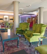Een stijlvolle lounge in het Hillyard House Hotel in Castlewellan heeft groenfluwelen fauteuils, paarse stoelen, kroonluchters en een open haard. Zuilen, een trap met rode vloerbedekking, moderne kunst en zachtpaarse verlichting creëren een chique, uitnodigende sfeer. | Echt Ierland Vakanties