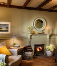 Gezellige woonkamer met houten balken, een brandende houtkachel en twee fauteuils, een beige met een geel kussen en een donkergrijze. Een rieten mand bevat houtblokken en kaarsen branden op een witte schoorsteenmantel onder een ronde spiegel. Bloemen en zachte verlichting creëren een warme, uitnodigende sfeer. | Echt Ierland Vakanties