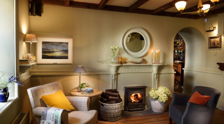 Gezellige woonkamer met houten balken, een brandende houtkachel en twee fauteuils, een beige met een geel kussen en een donkergrijze. Een rieten mand bevat houtblokken en kaarsen branden op een witte schoorsteenmantel onder een ronde spiegel. Bloemen en zachte verlichting creëren een warme, uitnodigende sfeer. | Echt Ierland Vakanties