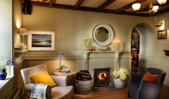 Gezellige woonkamer met houten balken, een brandende houtkachel en twee fauteuils, een beige met een geel kussen en een donkergrijze. Een rieten mand bevat houtblokken en kaarsen branden op een witte schoorsteenmantel onder een ronde spiegel. Bloemen en zachte verlichting creëren een warme, uitnodigende sfeer. | Echt Ierland Vakanties