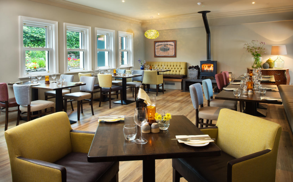 Licht, gezellig restaurant met netjes gedekte tafels en kleurrijke stoelen. Grote ramen laten daglicht binnen, bloemen en groen zijn buiten zichtbaar. Een houtkachel gloeit in de hoek. Muurkunst, vazen en een lamp geven de uitnodigende ruimte een huiselijk tintje. | Echt Ierland Vakanties