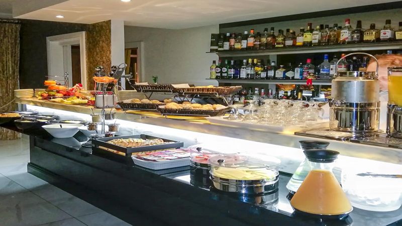 Op een buffettafel in het Brook Lodge Hotel in Killarney staan ontbijtproducten zoals fruit, gebak, kazen en sappen. Achter het buffet bevindt zich een bar met rekken met verschillende likeurflessen en glaswerk, in een moderne, georganiseerde ruimte. | Echt Ierland Vakanties