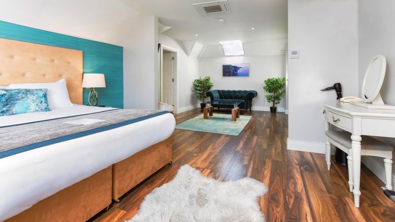 Een ruime, moderne slaapkamer in het Hillyard House Hotel in Castlewellan heeft houten vloeren, een groot bed met een gebruind hoofdeinde, een groenblauwe accentmuur, een donkergroene bank, een kaptafel met spiegel, een pluche wit vloerkleed, planten in potten en heldere natuurlijke verlichting. | Echt Ierland Vakanties