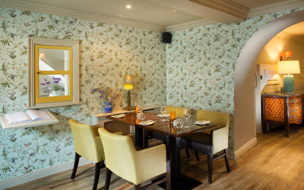 Een gezellige eethoek met een donkerhouten tafel voor vier personen, omringd door lichtgeel gestoffeerde stoelen. De muren zijn bedekt met gebloemd behang en er is een raam, een plank met boeken, een vaas met bloemen en warme verlichting in de hele kamer. | Echt Ierland Vakanties