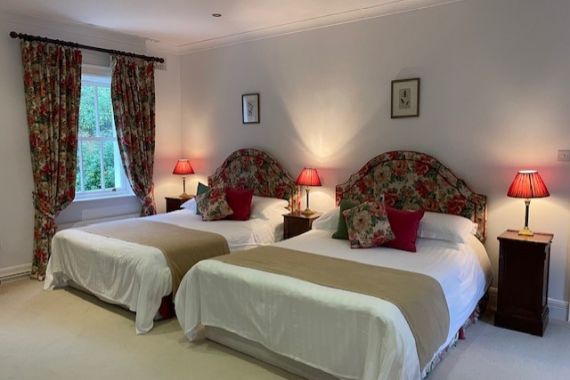 Een gezellige slaapkamer met twee tweepersoonsbedden, elk met een hoofdeinde met bloemmotief, rode en groene accentkussens en beige plaids. Bloemengordijnen omlijsten een raam en drie rode lampen staan op houten nachtkastjes. De muren en het tapijt zijn lichtgekleurd. | Echt Ierland Vakanties