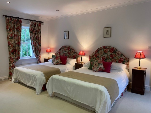 Een gezellige slaapkamer met twee tweepersoonsbedden, elk met een hoofdeinde met bloemmotief, rode en groene accentkussens en beige plaids. Bloemengordijnen omlijsten een raam en drie rode lampen staan op houten nachtkastjes. De muren en het tapijt zijn lichtgekleurd. | Echt Ierland Vakanties