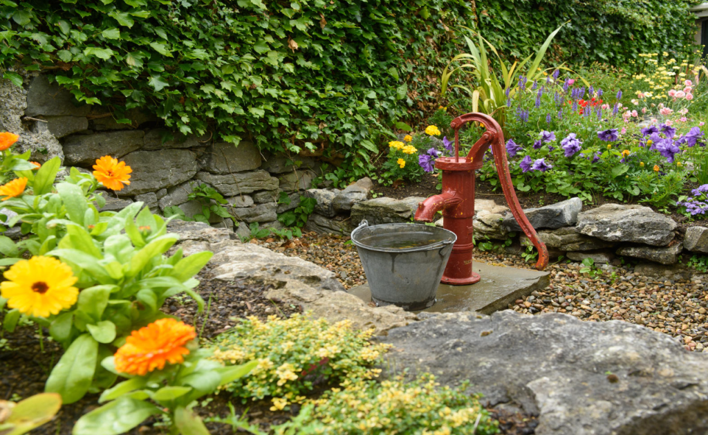 Een rode handwaterpomp met een grijze metalen emmer staat op een stenen voet in een weelderige tuin. Fel oranje en gele bloemen, groene planten en kleurrijke bloemen omringen de pomp. Een stenen muur bedekt met klimop vormt de achtergrond. Het tafereel oogt rustig en rustiek. | Echt Ierland Vakanties