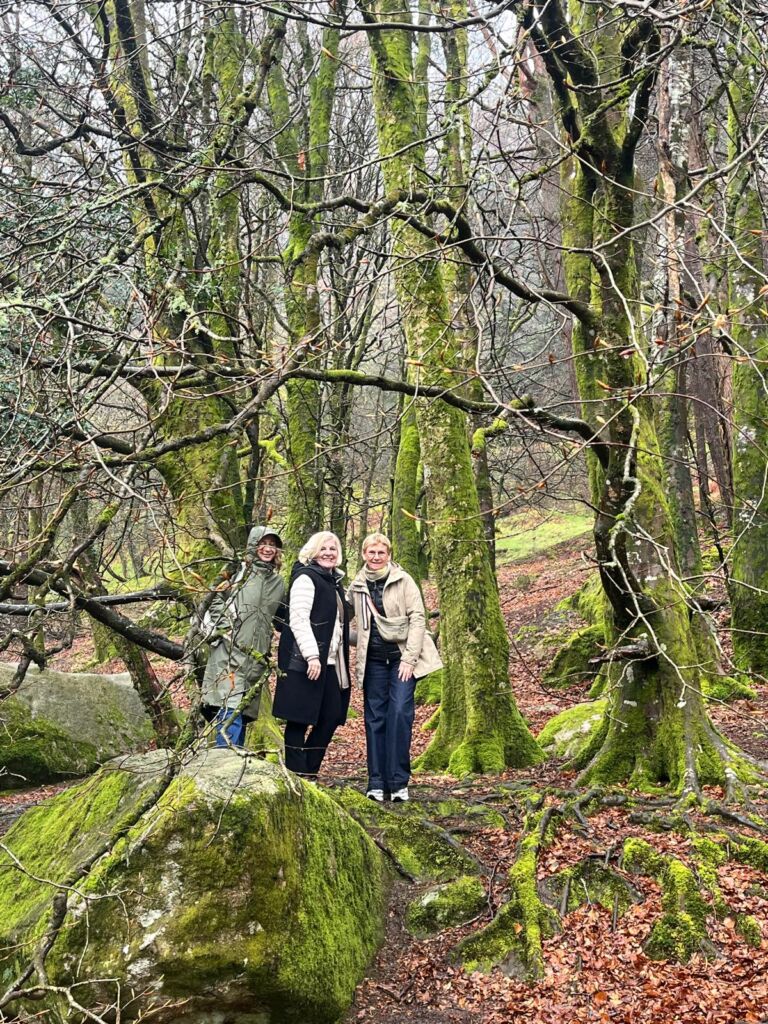 Drie mensen staan glimlachend in een bemost bos met kale bomen en verspreide rotsen. Ze dragen winterjassen en mutsen, omringd door levendig groen mos en gevallen bruine bladeren, wat duidt op een koele, vochtige dag. De stemming is vrolijk en ontspannen. | Echt Ierland Vakanties