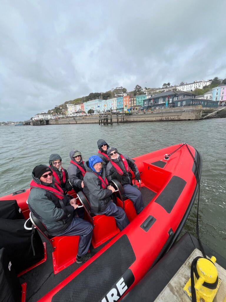 Zes mensen, warm gekleed en met rode reddingsvesten aan, zitten in een felrode opblaasboot op het water. Achter hen staat een rij kleurrijke gebouwen aan de waterkant onder een bewolkte hemel. De groep lijkt klaar voor een uitstapje of rondleiding. | Echt Ierland Vakanties