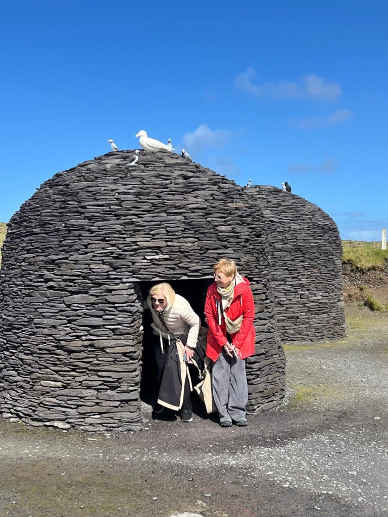 Twee vrouwen verlaten een kleine, koepelvormige stenen hut onder een strakblauwe hemel. De ene vrouw, in zwart en beige, hurkt bij de ingang, terwijl de andere, in een rode jas, naast haar staat. Bovenop de stenen structuur zitten verschillende witte vogels. | Echt Ierland Vakanties