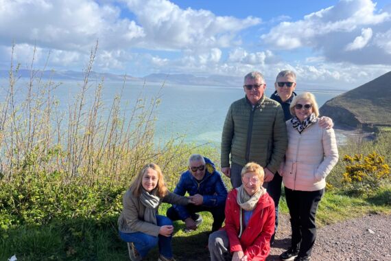 Zes volwassenen poseren glimlachend op een pad aan zee met blauwe lucht en wolken boven hun hoofd. Vier staan terwijl twee voorover hurken. Struiken en de oceaan zijn op de achtergrond en bergen in de verte steken boven het water uit. Iedereen draagt jassen en zonnebrillen. | Echt Ierland Vakanties