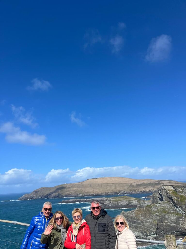 Vijf volwassenen staan glimlachend voor een schilderachtig kustlandschap met ruige kliffen en de oceaan. De lucht is helderblauw met een paar wolken. Ze dragen winterjassen, zonnebrillen en zien er vrolijk uit. Heuvels en golven zijn zichtbaar op de achtergrond. | Echt Ierland Vakanties