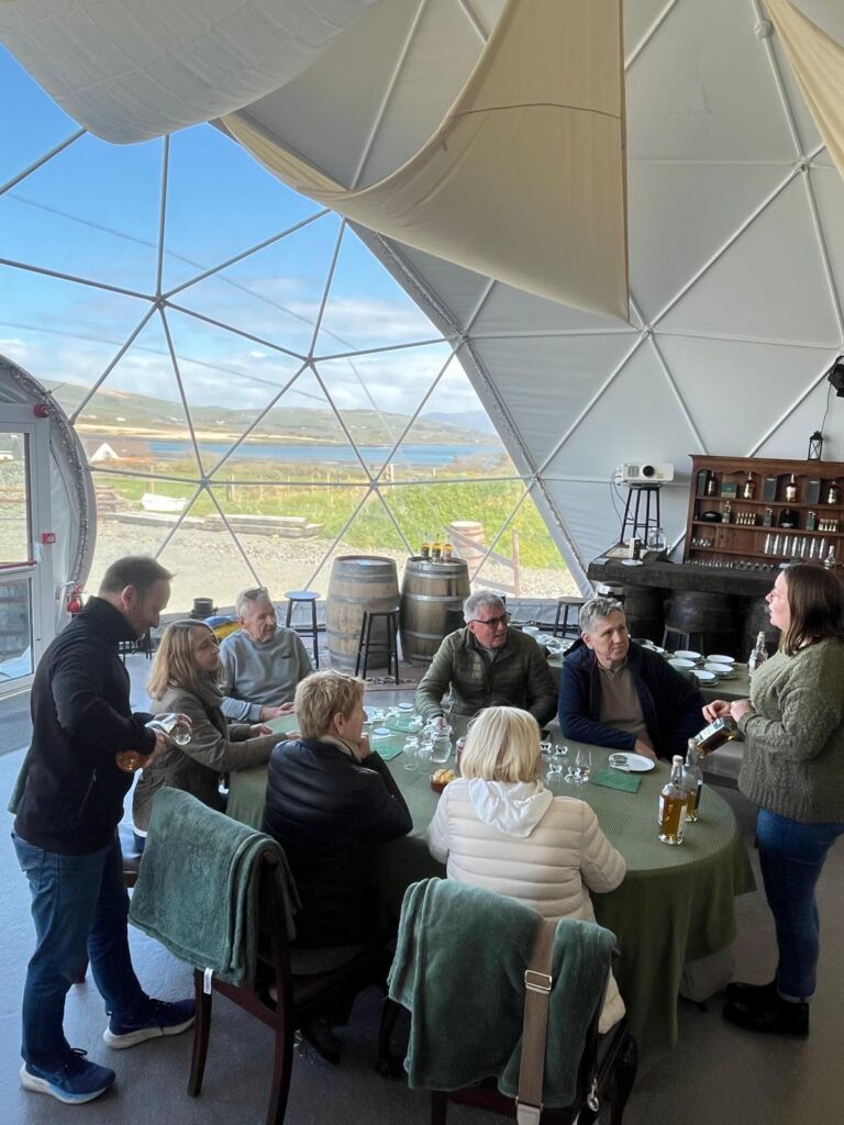 Een groep van acht volwassenen zit en staat rond een ronde tafel in een heldere geodetische koepel. Een vrouw schenkt drankjes in terwijl de anderen toekijken. Grote ramen onthullen een schilderachtig landschap met heuvels, water en blauwe lucht buiten. Planken en vaten versieren het interieur. | Echt Ierland Vakanties