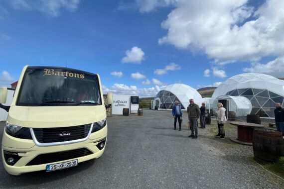Een crèmekleurige tourbus van Bartons staat geparkeerd op grind in de buurt van drie witte geodetische koepels. Verschillende mensen staan en lopen rond het gebied onder een gedeeltelijk bewolkte hemel. Houten vaten en een wit bord met de tekst "Food Voyage" zijn zichtbaar op de achtergrond. | Echt Ierland Vakanties