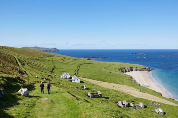 Twee mensen lopen een met gras begroeide heuvel af in de richting van witte huisjes bij een zandstrand. Glooiende groene velden, stenen hekken en verspreide rotsen leiden naar de blauwe zee onder een heldere hemel, met verre eilanden zichtbaar aan de horizon. | Echt Ierland Vakanties