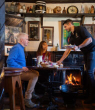 Een ober serveert eten aan een man en vrouw aan een houten tafel in een gezellige, rustieke pub met donkerhouten interieur, een brandende open haard, ingelijste foto's en antieke voorwerpen op planken. De sfeer is warm en uitnodigend. | Echt Ierland Vakanties