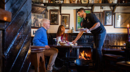 Een ober serveert eten aan een man en vrouw aan een houten tafel in een gezellige, rustieke pub met donkerhouten interieur, een brandende open haard, ingelijste foto's en antieke voorwerpen op planken. De sfeer is warm en uitnodigend. | Echt Ierland Vakanties