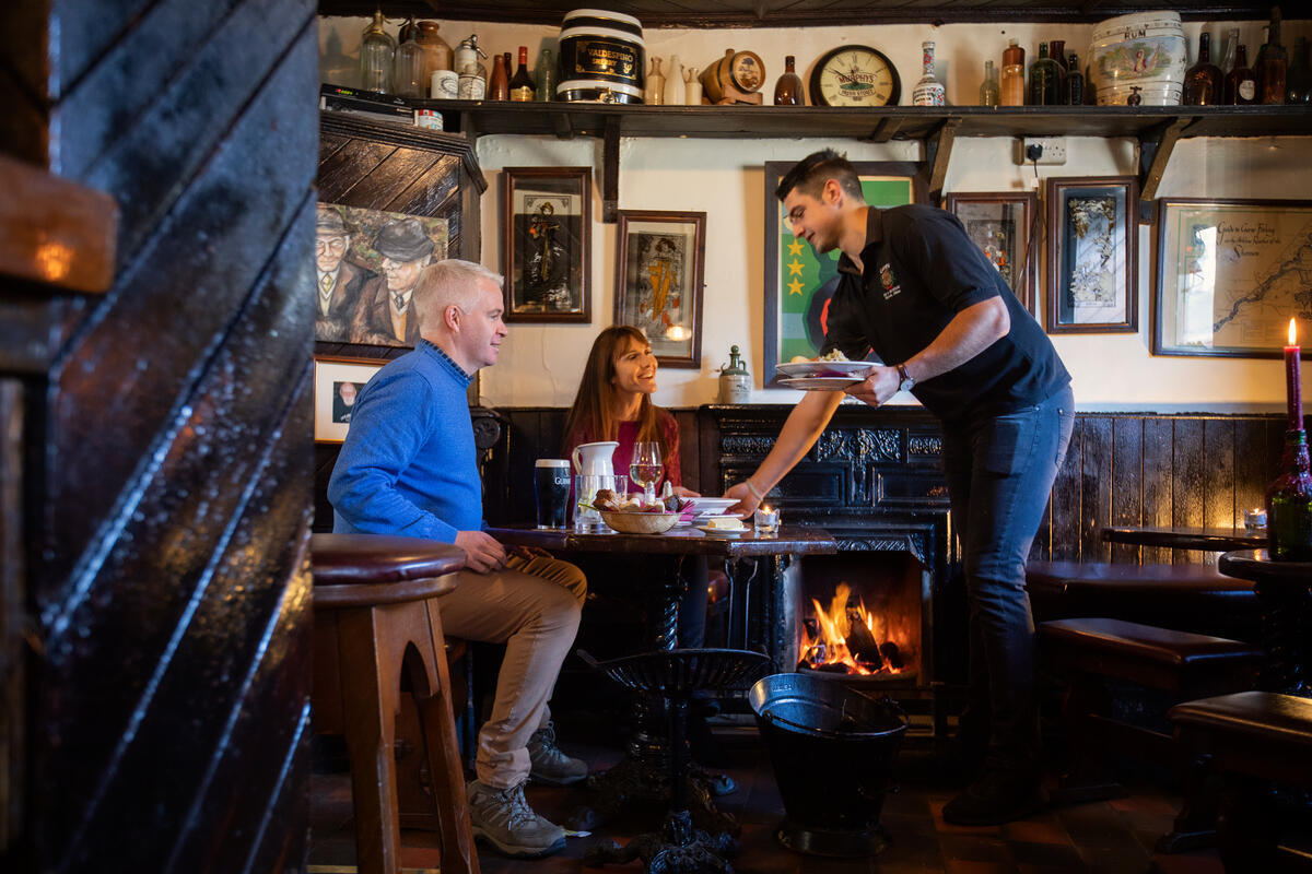 Een ober serveert eten aan een man en vrouw aan een houten tafel in een gezellige, rustieke pub met donkerhouten interieur, een brandende open haard, ingelijste foto's en antieke voorwerpen op planken. De sfeer is warm en uitnodigend. | Echt Ierland Vakanties