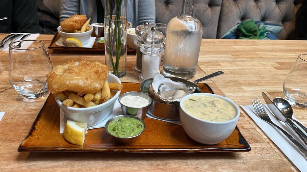 Een houten tafel gedekt voor een maaltijd bevat een dienblad met gebakken vis en friet, erwten, tartaarsaus, een citroenpartje, een kom romige vissoep en een schaal met oesters op ijs. Glazen, bestek, zout, peper en een waterkaraf staan ook op tafel. | Echt Ierland Vakanties
