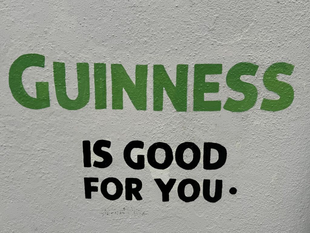 Tekst geschilderd op een ruwe witte muur luidt: GUINNESS in grote groene letters, met IS GOED VOOR JE. eronder in vette zwarte letters. De tekst is gecentreerd en de muurtextuur is zichtbaar in de hele afbeelding. | Echt Ierland Vakanties