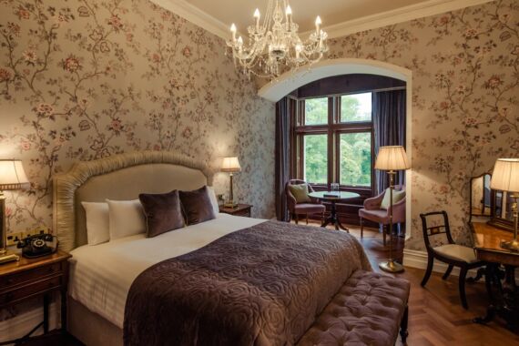 Elegante slaapkamer met bloemetjesbehang, een groot gestoffeerd bed met paars beddengoed, nachtkastjes met lampjes, een kristallen kroonluchter, een raam met gordijnen, twee fauteuils met een tafeltje en een bankje aan het voeteneind van het bed. | Echt Ierland Vakanties
