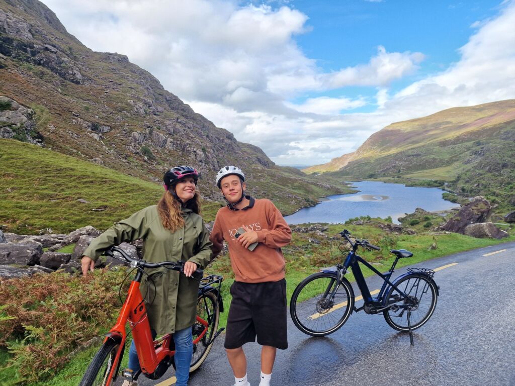 Twee lachende mensen met helmen staan met fietsen op een bergweg. Groene heuvels en een kronkelende blauwe rivier strekken zich uit in de verte onder een gedeeltelijk bewolkte hemel, waardoor een schilderachtig en vredig landschap ontstaat. De weg maakt zachte bochten langs het water. | Echt Ierland Vakanties
