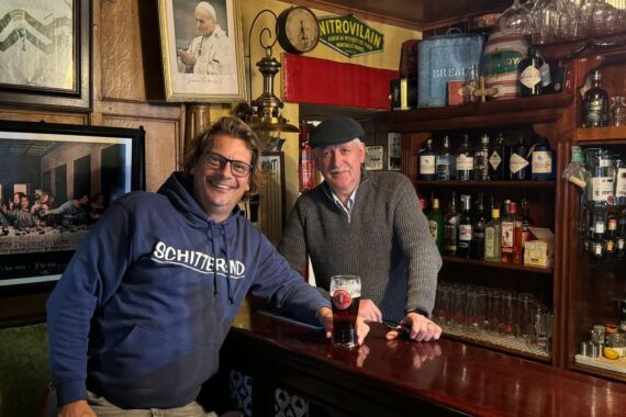 Twee lachende mannen poseren in een gezellig café. De ene, in een blauwe hoodie, leunt op de bar met een drankje. De andere, in een grijze trui en platte pet, staat achter de bar. Planken met flessen, glazen, ingelijste foto's en vintage borden vullen de warm verlichte achtergrond. | Echt Ierland Vakanties