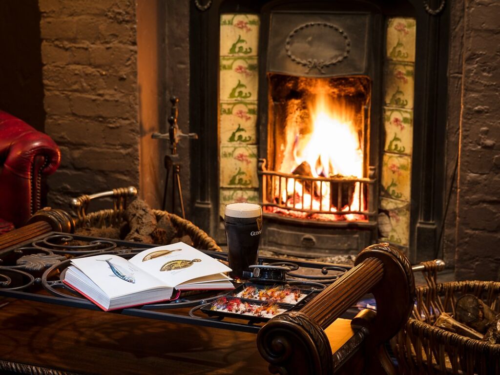 Een gezellige kamer met een knetterende open haard; een pint donker bier, een aquarelverfset en een open boek met geïllustreerde vissen liggen op een houten tafel. Een roodleren stoel en rieten manden gevuld met brandhout staan vlakbij. Een warme, uitnodigende sfeer. | Echt Ierland Vakanties