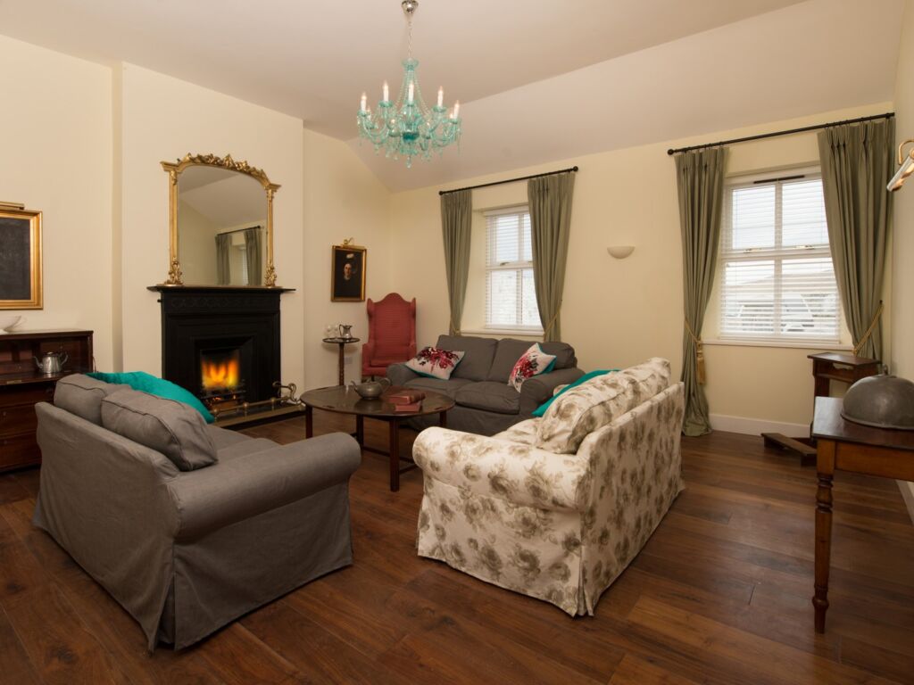Een gezellige woonkamer met houten vloeren, twee grijze banken en een gebloemde bank rond een houten salontafel, een open haard met een goudomrande spiegel erboven, een turkooizen kroonluchter, grote ramen met groene gordijnen en klassieke meubels. | Echt Ierland Vakanties