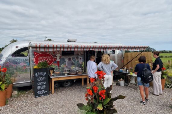 Een zilverkleurige foodtruck met een rood-witte luifel bedient klanten in de open lucht. Vier mensen staan in de rij bij een menubord. Rode bloemen en potplanten sieren het grindterrein, en op de achtergrond strekken groene velden zich uit onder een bewolkte hemel. | Echt Ierland Vakanties