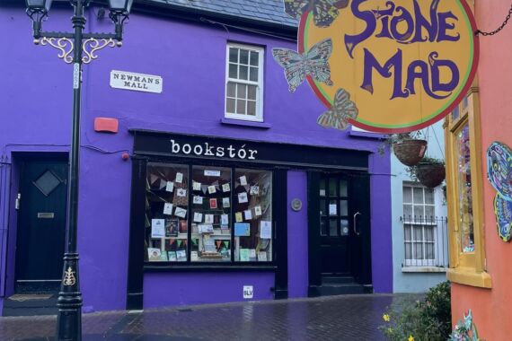 Een kleurrijk straatbeeld toont een paarse boekhandel met boeken in de etalage, een zwarte deur en een bordje met het opschrift „bookstór“. Vlakbij hangt een geel „Stone Mad“-bord met vlinders. De stoep is nat en de gebouwen zijn in levendige kleuren geschilderd. | Echt Ierland Vakanties