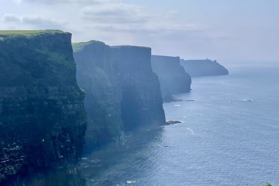 Een mistig uitzicht op de Cliffs of Moher in Ierland, met indrukwekkende, steile rotswanden die boven de blauwe Atlantische Oceaan uittorenen. De lucht is gedeeltelijk bewolkt en de toppen van de kliffen zijn bedekt met weelderig groen gras. | Echt Ierland Vakanties