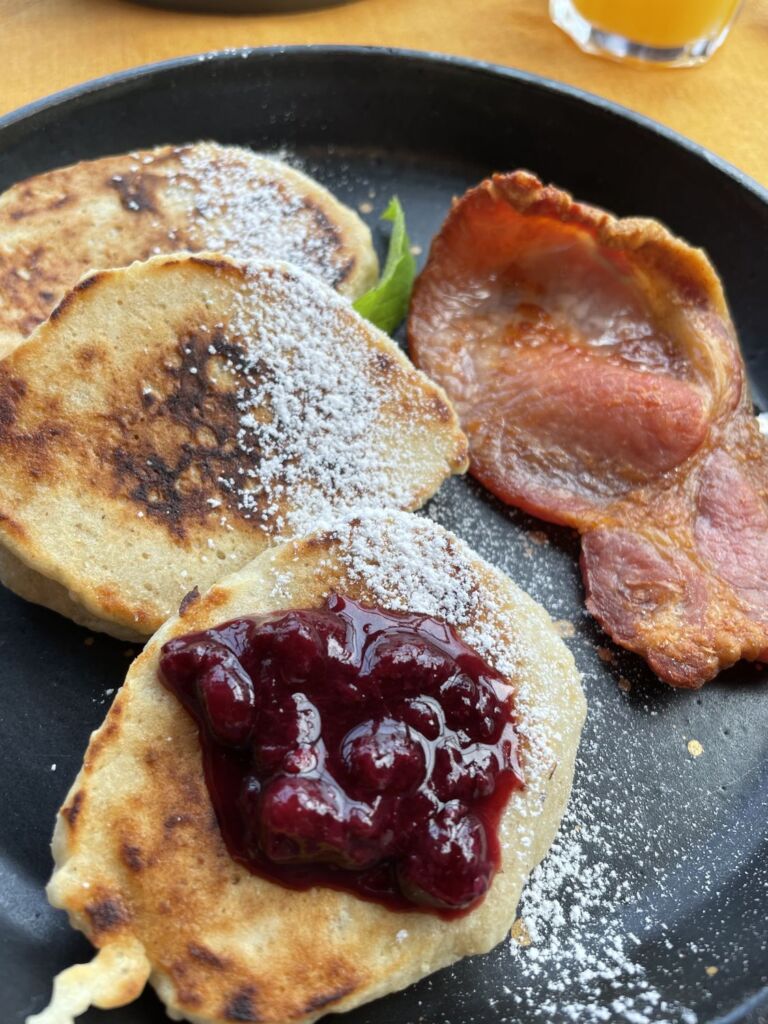 Op een zwart bord liggen drie dikke pannenkoeken, bestrooid met poedersuiker; op één ervan ligt een laagje compote van rode bessen. Naast de pannenkoeken liggen twee stukjes knapperig gebakken spek, en daartussen is een klein groen muntblaadje te zien. | Echt Ierland Vakanties