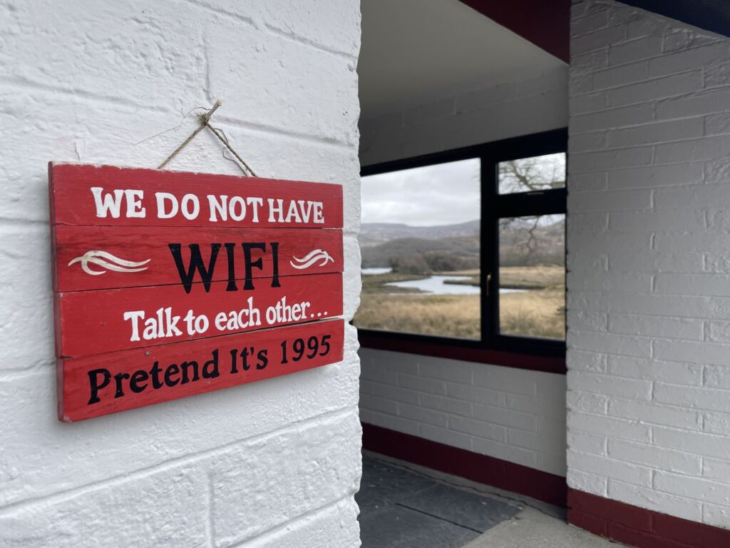 Op een rood-wit houten bord aan een witte bakstenen muur staat: „WIJ HEBBEN GEEN WIFI. Praat met elkaar... Doe alsof het 1995 is.” Op de achtergrond is een raam te zien met een prachtig uitzicht op velden, bomen en een rivier. | Echt Ierland Vakanties