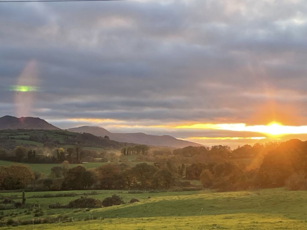 Een schilderachtig landschap bij zonsondergang, met glooiende groene heuvels, hier en daar een boom en velden. De zon zakt achter de horizon en werpt een gouden gloed, terwijl wolken delen van de hemel bedekken. In de verte tekenen de heuvels zich af tegen de kleurrijke hemel. | Echt Ierland Vakanties