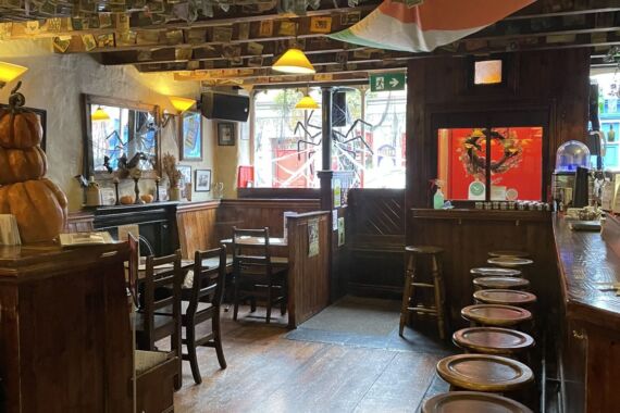 Een gezellige pub met houten tafels en krukken, een bar aan de rechterkant en dollarbiljetten die aan het plafond zijn geprikt. Warme verlichting, ingelijste foto’s en een Ierse vlag sieren de ruimte. Zonlicht schijnt door een raam en een open deur aan de achterkant. | Echt Ierland Vakanties