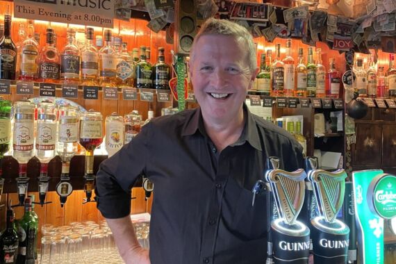 Een barman met kort haar en een snor glimlacht achter de bar, omringd door flessen en Guinness-tapkranen. Boven hem hangt een bordje met de tekst „Live Irish Music”, terwijl er glazen boven zijn hoofd hangen. De gezellige bar is sfeervol verlicht, met zichtbare planken vol sterke drank en biertapkranen. | Echt Ierland Vakanties